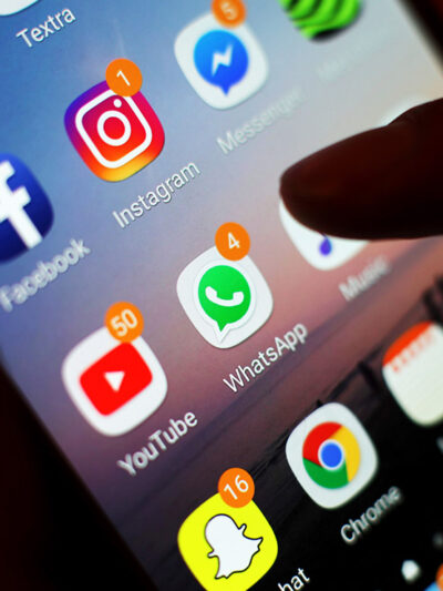 A hand holding a phone whose screen displays a group of apps, including Facebook, Instagram, and WhatsApp.