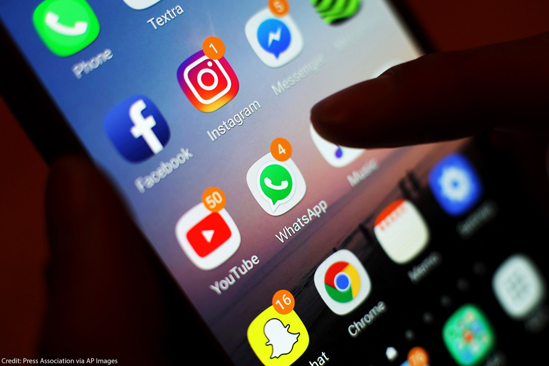 A hand holding a phone whose screen displays a group of apps, including Facebook, Instagram, and WhatsApp.