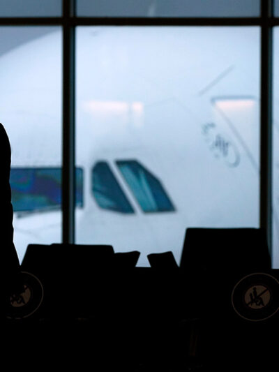 The silhouette of a pilot wearing a facemask walking past a window with a Delta plane outside.