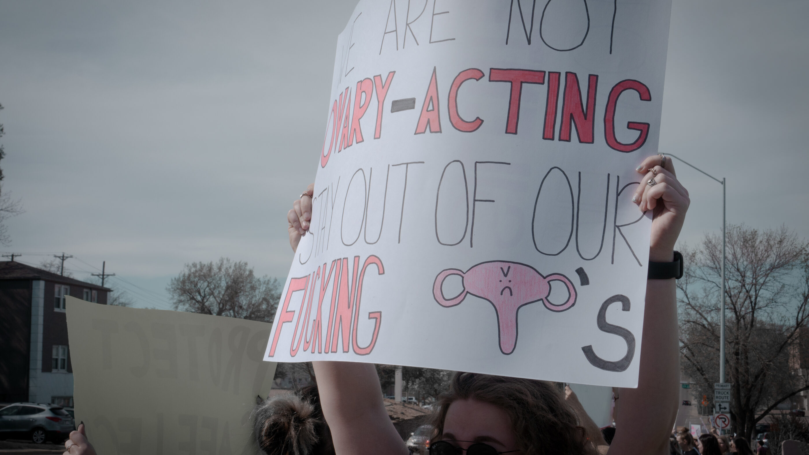 Image of a person holding a protest sign that reads, "We are not ovary-acting. Keep out of our fucking uteruses."