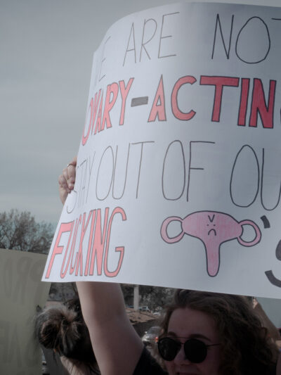 Image of a person holding a protest sign that reads, "We are not ovary-acting. Keep out of our fucking uteruses."