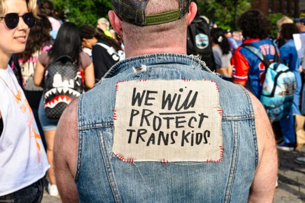 At a trans rally, person (wearing a denim vest and with their back to the camera) displays the sign stitched to the vest that reads "WE WILL PROTECT TRANS KIDS."