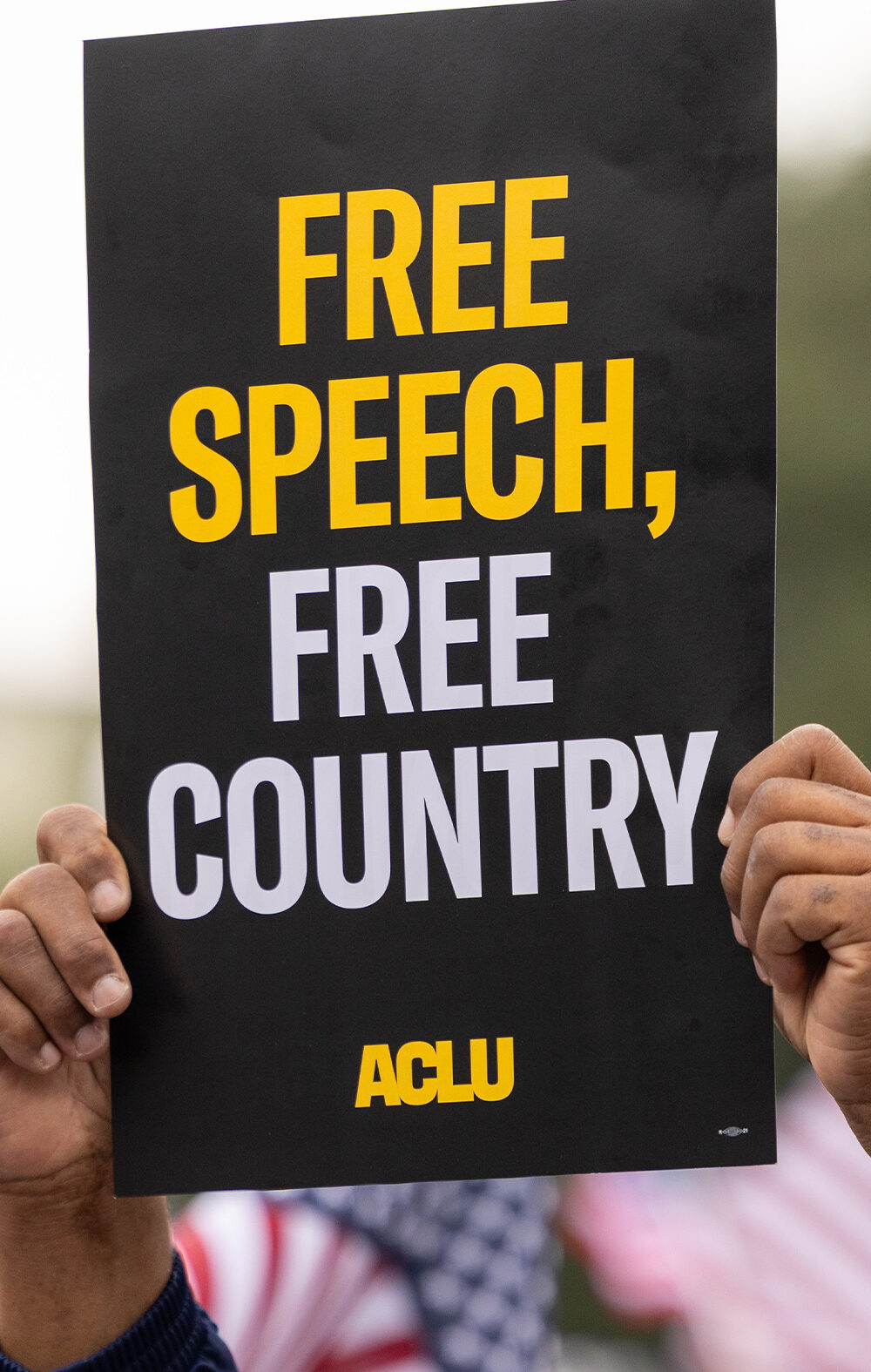 A demonstrator (with only their hands visible) holds up a sign that reads " FREE SPEECH, FREE COUNTRY - ACLU".