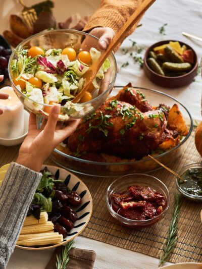 A multi-ethnic group of people sharing a festive, communal meal at a dining table with roasted chicken, salad, bread, fruit, and wine.