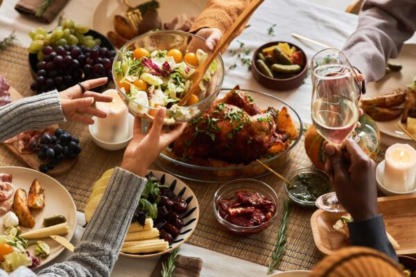 A multi-ethnic group of people sharing a festive, communal meal at a dining table with roasted chicken, salad, bread, fruit, and wine.