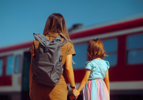 A photo of a woman wearing a tan dress and a backpack holding the hand of a child with red hear and a pink and blue dress.