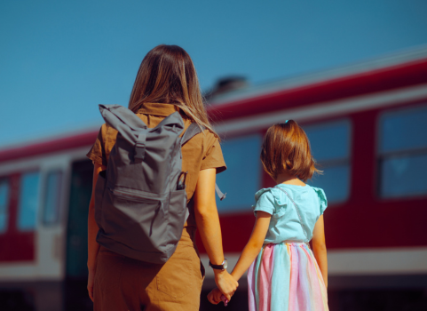A photo of a woman wearing a tan dress and a backpack holding the hand of a child with red hear and a pink and blue dress.