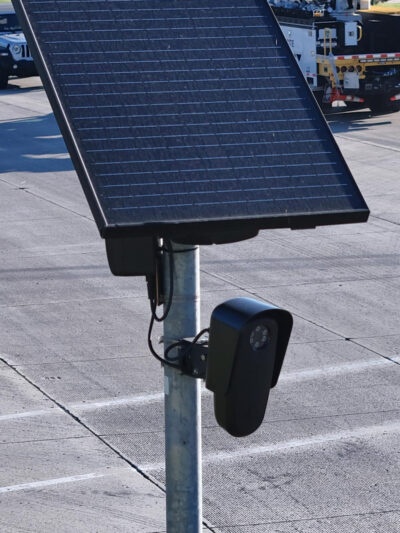 A solar-powered Flock camera is mounted on a pole beside a busy multi-lane road, with cars and a utility truck driving through the intersection in the background.