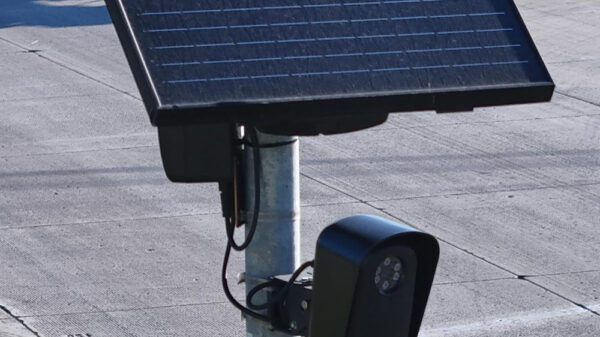 A solar-powered Flock camera is mounted on a pole beside a busy multi-lane road, with cars and a utility truck driving through the intersection in the background.