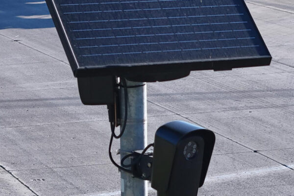 A solar-powered Flock camera is mounted on a pole beside a busy multi-lane road, with cars and a utility truck driving through the intersection in the background.
