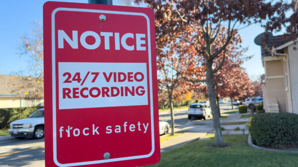 A red Flock Safety sign on a pole reads "Notice: 24/7 Video Recording" on a residential street, with parked cars, homes, trees, and a clear blue sky in the background.